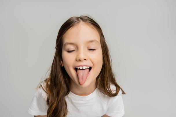 Preteen kid sticking out tongue and closing eyes isolated on grey   - Foto, Imagen