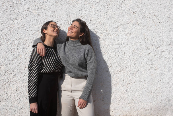 Portrait two girls couple in their twenties looking sky. Clothes with gray, white and black tones. White stuccoed wall background - Fotografie, Obrázek