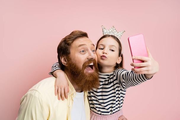 Preteen girl with crown headband pouting lips and taking selfie with bearded dad isolated on pink   - Φωτογραφία, εικόνα