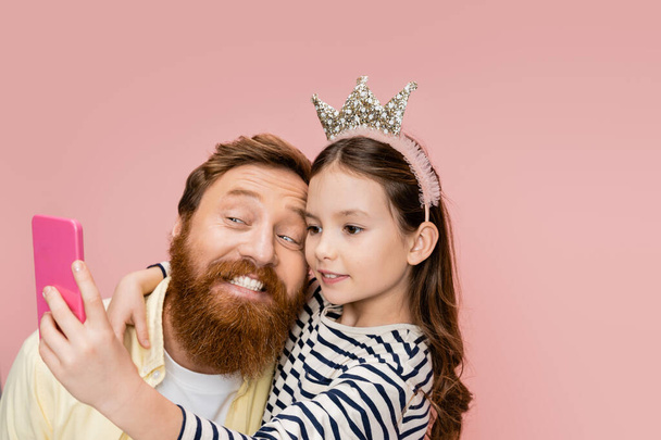 Preteen girl with crown headband taking selfie with cheerful father isolated on pink   - Fotoğraf, Görsel