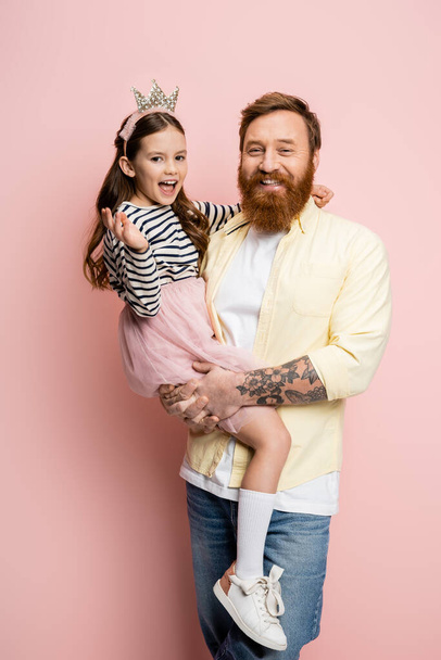 Cheerful bearded man holding preteen daughter in crown headband on pink background  - Fotó, kép