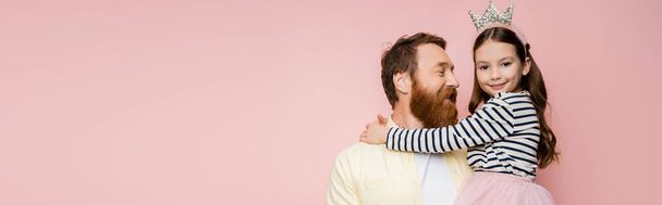 Smiling bearded man looking at preteen daughter with crown headband isolated on pink, banner  - 写真・画像