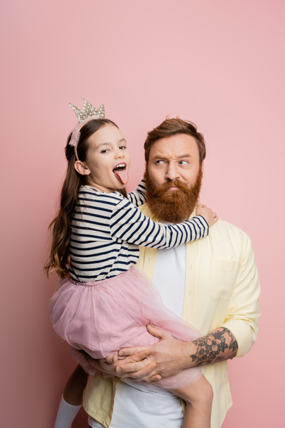 Tattooed father holding preteen daughter in crown headband sticking out tongue on pink background  - Φωτογραφία, εικόνα