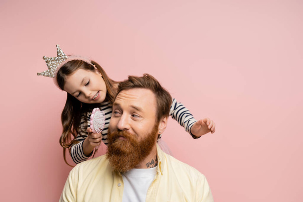 Sonriente chica con corona diadema celebración cepillo cerca barbudo padre aislado en rosa   - Foto, Imagen