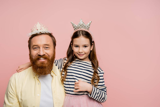 Усміхнена дочка і батько в коронних головних уборах дивиться на камеру ізольовано на рожевому
   - Фото, зображення