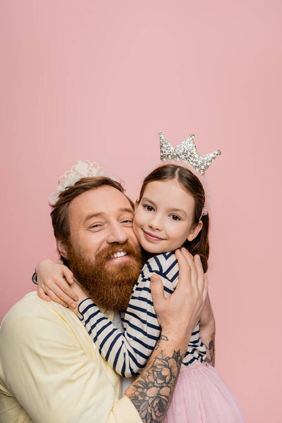 Sonriente hombre tatuado en diadema de corona abrazando hija aislada en rosa   - Foto, imagen