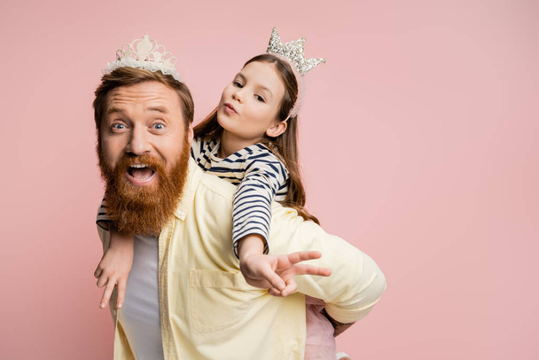 Preteen kid in crown headband showing peace sign while piggybacking on dad isolated on pink   - Фото, изображение
