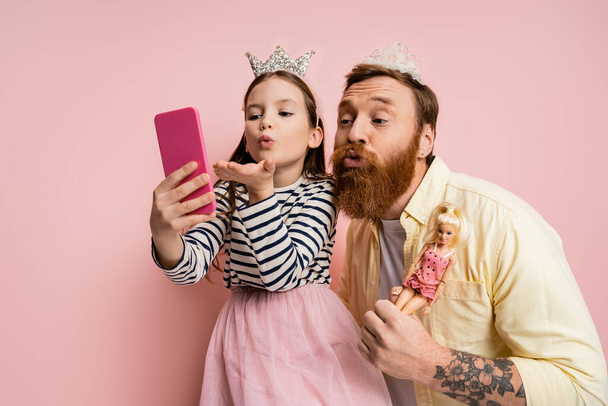 Daughter and father with doll and crown headbands taking selfie on smartphone on pink background  - Foto, imagen