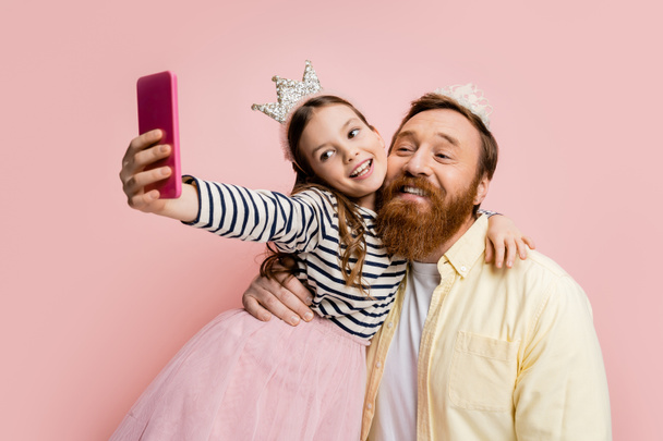 Smiling girl hugging father in crown headband and taking selfie isolated on pink   - Фото, изображение