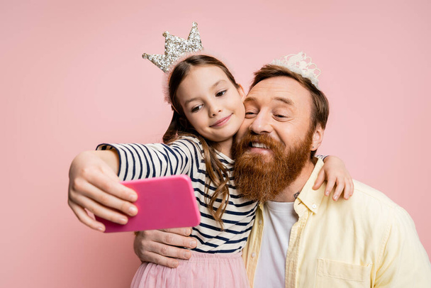 Positive man with crown headband taking selfie with daughter isolated on pink   - Фото, изображение