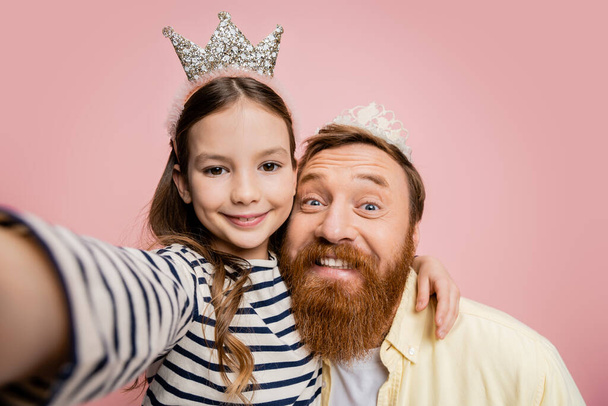 Enfant souriant étreignant père avec bandeau couronne et regardant la caméra isolée sur rose   - Photo, image