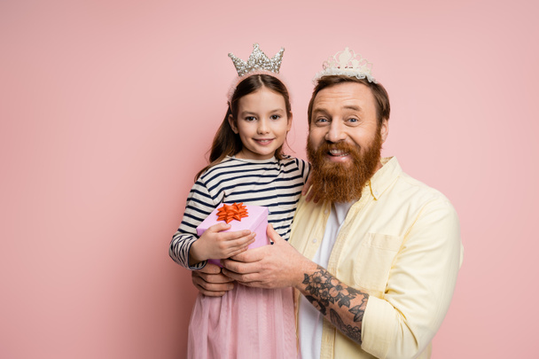 Tattooed man with crown headband holding gift near preteen daughter on pink background  - Фото, зображення