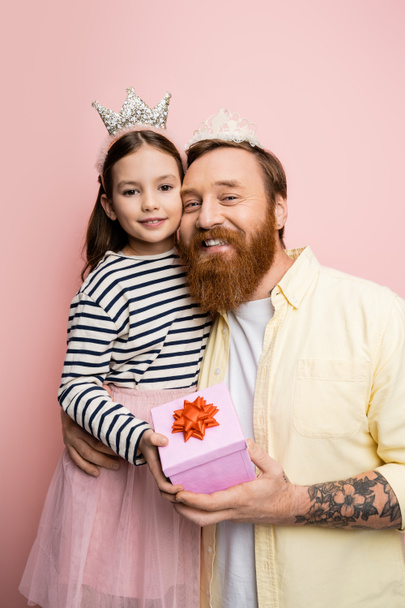 Cheerful man and child in crown headbands holding present on pink background  - 写真・画像