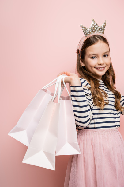 Cheerful preteen kid in crown headband holding shopping bags and looking at camera on pink background - Fotoğraf, Görsel