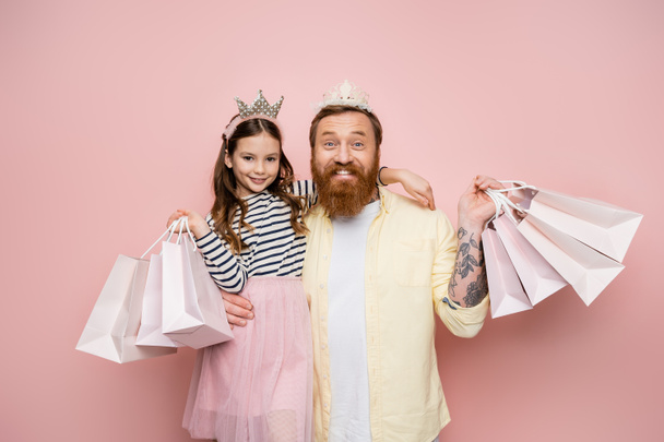 Cheerful man and daughter with crown headbands holding shopping bags on pink background  - Фото, изображение