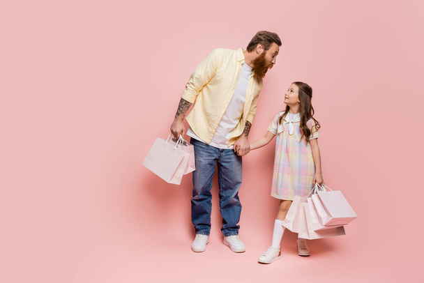 Tattooed man holding hand of daughter with purchases on pink background  - Photo, Image