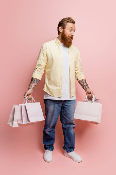 Confused tattooed man holding shopping bags on pink background - Foto, immagini