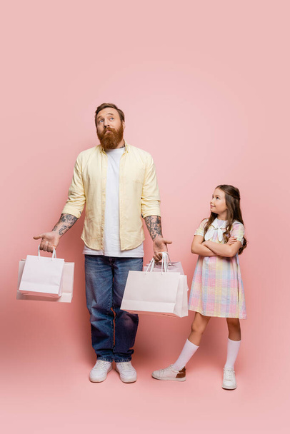 Chica preadolescente cruzando brazos cerca de padre pensativo con bolsas de compras sobre fondo rosa  - Foto, Imagen