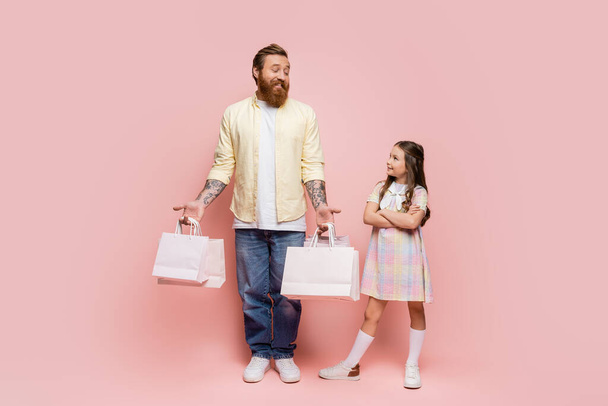 Teljes hossza lány keresztbe karok közelében mosolygós apa bevásárló táskák rózsaszín háttér  - Fotó, kép