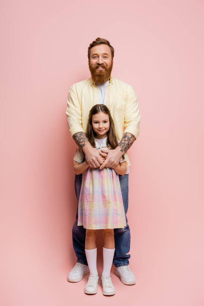 Полная длина улыбающийся татуированный мужчина обнимает дочь на розовом фоне  - Фото, изображение