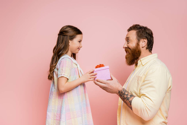 side view of cheerful bearded man giving present to daughter isolated on pink   - 写真・画像