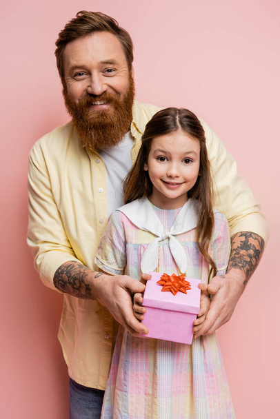 Θετικός άντρας με τατουάζ που κρατάει κουτί δώρου κοντά στην κόρη του σε ροζ φόντο  - Φωτογραφία, εικόνα