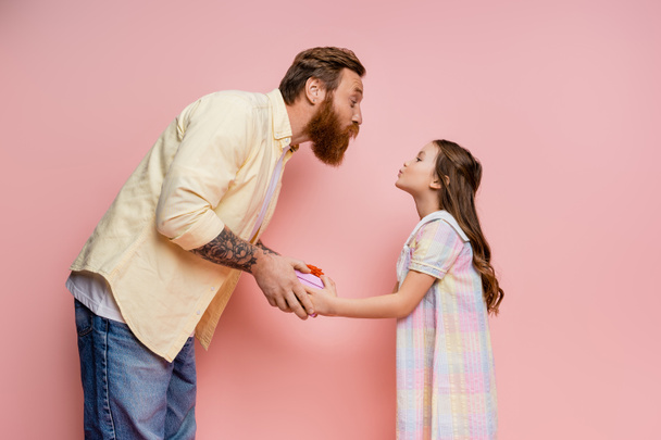 Side view of girl pouting lips while giving gift to bearded dad on pink background  - Фото, изображение