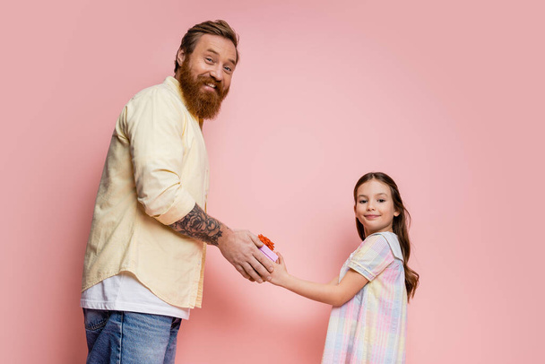 Vrolijke baard vader en tiener meisje houden geschenkdoos en kijken naar de camera op roze achtergrond  - Foto, afbeelding