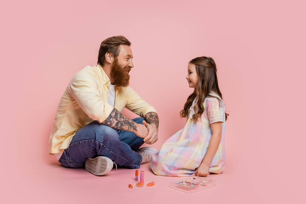 Side view of smiling tattooed father sitting near daughter and decorative cosmetics on pink background  - Photo, Image