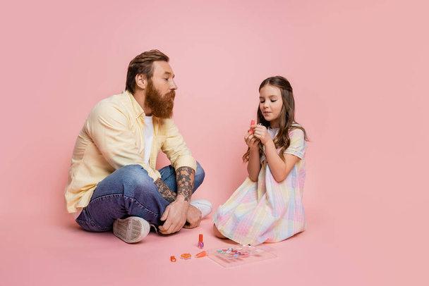 Preteen girl holding lipstick near tattooed dad on pink background  - Photo, Image
