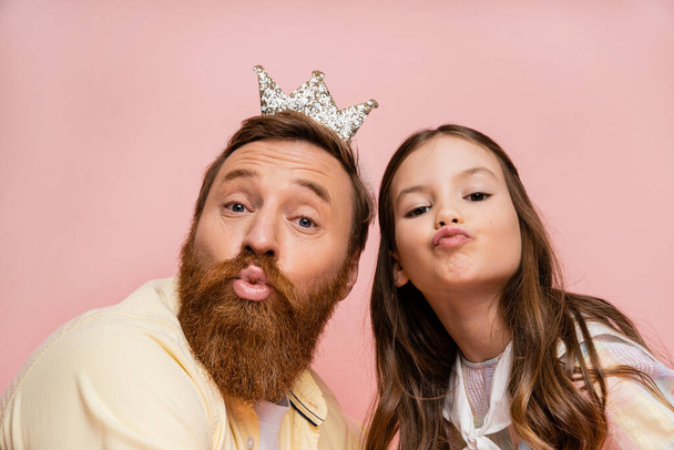 Bearded dad with crown headband pouting lips near daughter isolated on pink   - 写真・画像