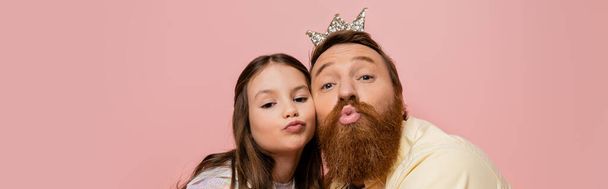 Bearded man with crown headband pouting lips near daughter isolated on pink, banner  - Foto, Imagem