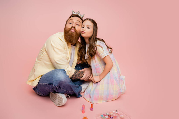 Bärtiger Mann mit Krone Haarband schmollen Lippen in der Nähe der Tochter und dekorative Kosmetik auf rosa Hintergrund  - Foto, Bild