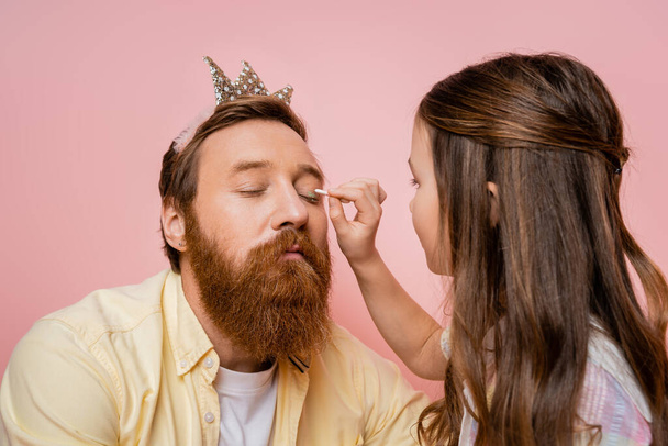 Preteen girl applying eyeshadow on father with crown headband isolated on pink   - Foto, Imagem