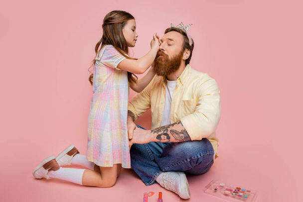 Preteen girl applying decorative cosmetic on tattooed dad with crown headband on pink background  - Foto, Bild