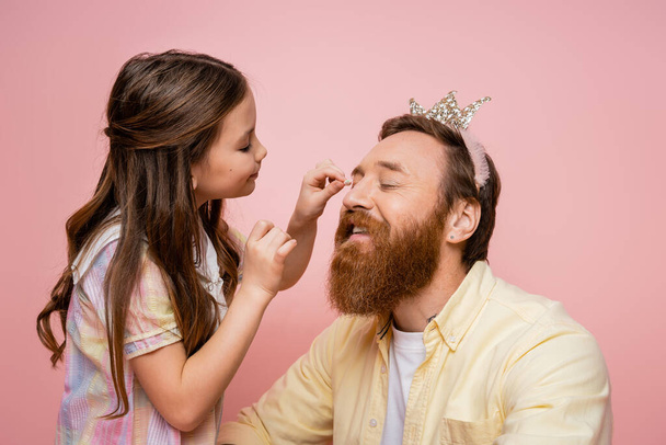 Smiling preteen girl applying eyeshadow on bearded dad with crown headband isolated on pink   - Фото, зображення