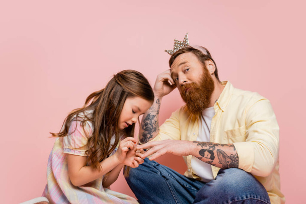 Preteen girl applying nail polish on hand of tattooed dad with crown headband on pink background  - Foto, Bild