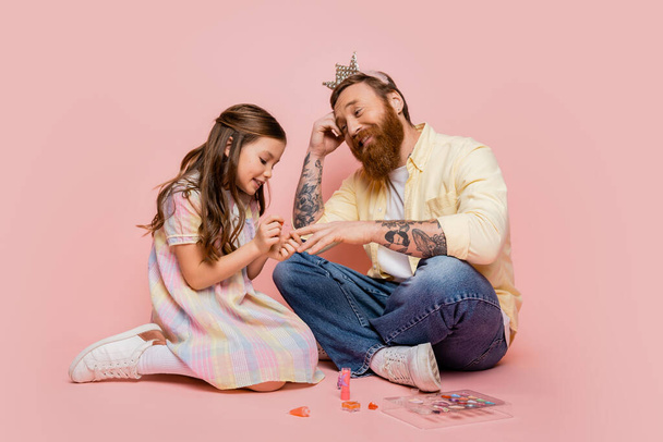 Cheerful girl applying nail polish on hand of tattooed dad with crown headband on pink background  - Фото, изображение