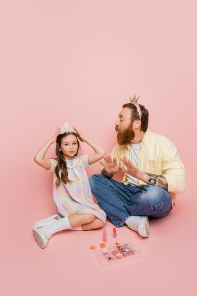 Szakállas apa koronával a fején nézi tizenéves lánya közelében dekoratív kozmetikumok rózsaszín háttér  - Fotó, kép