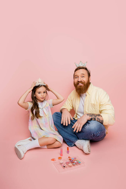 Позитивный бородатый мужчина смотрит в камеру рядом с дочерью с короной и декоративной косметикой на розовом фоне  - Фото, изображение
