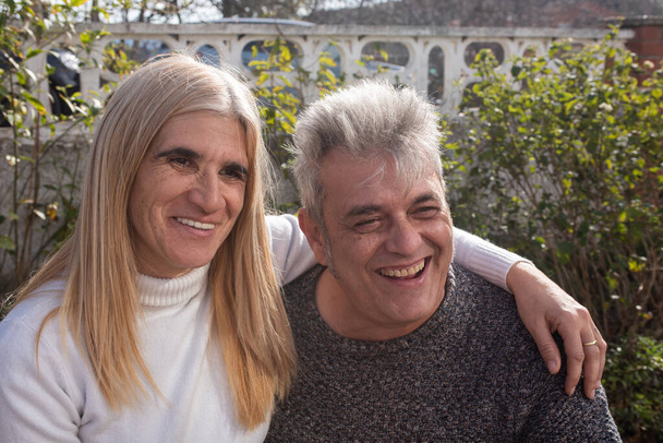 coppia di cinquantenni che si stringono a vicenda parlando e sorridendo, uomo dai capelli grigi in maglione grigio, donna con i capelli biondi tinti e maglione bianco - Foto, immagini