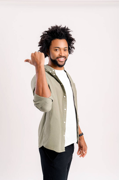 Vertical view of the joyful curly man presenting advertising area with big finger, holding empty place for commercial. Indoor studio shot isolated on white background - Фото, изображение