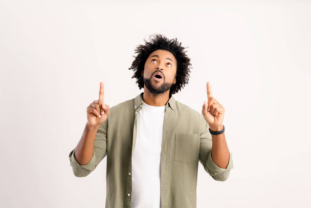 Advertising concept. Surprised African-American guy in shirt points fingers up isolated on white. Astonished multiracial man paying your attention at copy space, best deal, sale - Zdjęcie, obraz