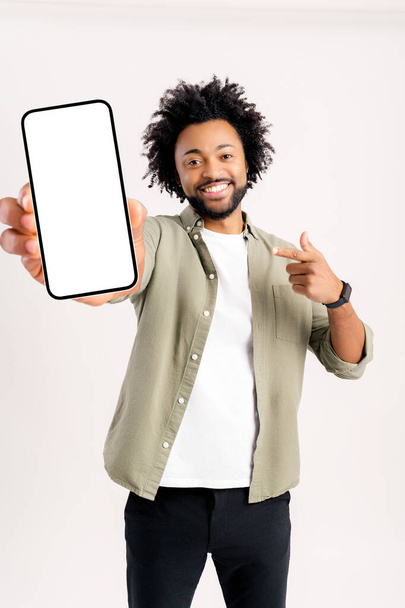 Portrait of satisfied brunette man with curly hair pointing at cellphone and smiling at camera, recommending gadget or mobile application. Indoor studio shot isolated on white background - Photo, image