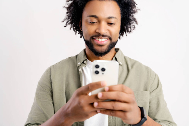 Portrait of cheerful young black guy sending message on mobile phone over white background. Happy African-American man using smartphone, enjoying chatting online, messaging in social networks - Fotó, kép