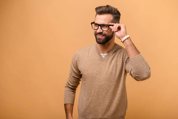 Attractive man isolated on beige background, guy standing and looks aside with friendly smile, male entrepreneur in casual wear adjusting stylish eyeglasses - Фото, изображение