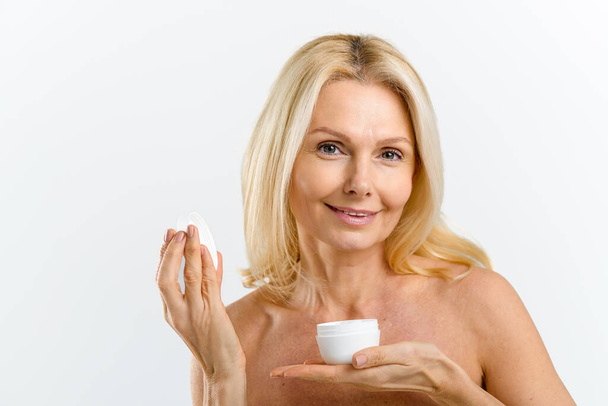 Sensitive middle-aged woman holding jar of moisturiser rich cream in hand isolated on white, inhales its fragrance with eyes closed, testing and advertising new cosmetology product - Fotoğraf, Görsel