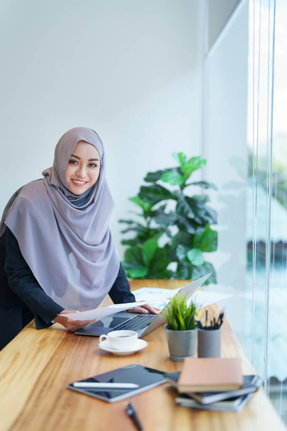 Eine schöne Muslimin zeigt morgens ein lächelndes Gesicht, indem sie Computer und Dokumente benutzt, die im Büro arbeiten. - Foto, Bild