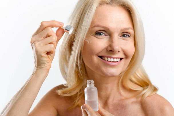 Close-up portrait of magnificent middle-aged blonde woman using skin care product, charming lady with radiant smooth face skin pipette of anti-aging serum smiling isolated on white - Fotografie, Obrázek