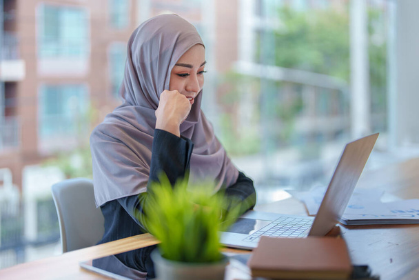 Egy gyönyörű muszlim nő, aki reggel mosolygós arcot vág az irodában dolgozó számítógépek és dokumentumok segítségével.. - Fotó, kép